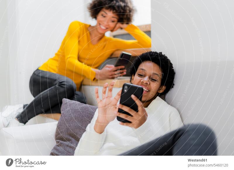 Two young women relaxing at home using their smartphones friends mate female friend couches settee settees sofa sofas telecommunication telephone telephones