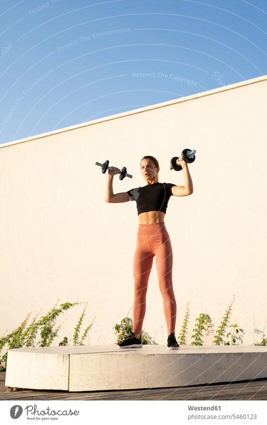 Sportswoman flexing muscle with dumbbell standing on pedestal against wall during sunrise color image colour image outdoors location shots outdoor shot