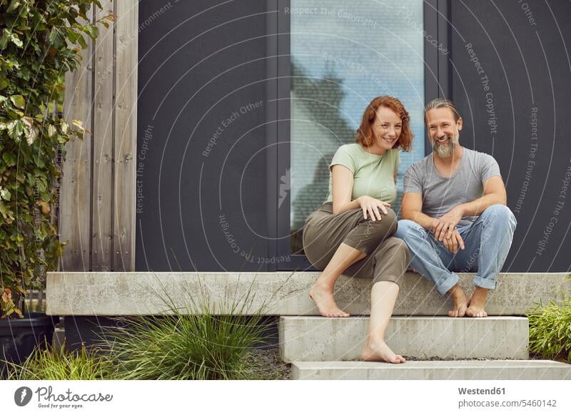 Smiling couple sitting on steps outside tiny house color image colour image Germany leisure activity leisure activities free time leisure time casual clothing