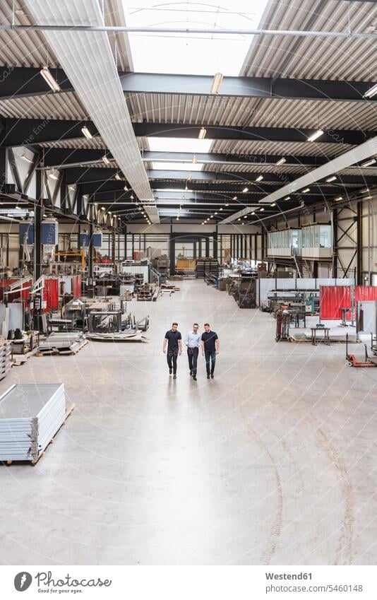 Three men walking and talking on factory shop floor speaking production hall colleagues man males going factories Adults grown-ups grownups adult people persons
