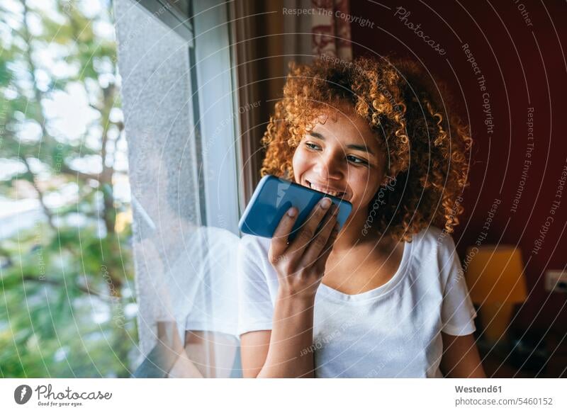 Young woman with curly hair sending voice message with cell phone next to the window voice mail voicemail voice messaging voice mails voicemails curls curled