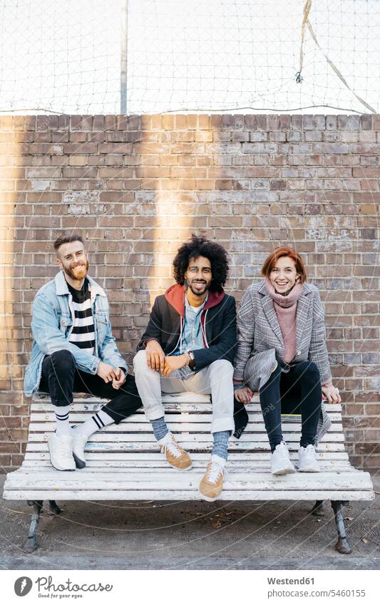 Portrait of three happy friends sitting on a bench in front of a brick wall brick walls benches Seated portrait portraits happiness friendship fashionable