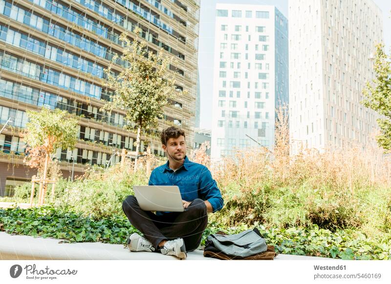 Businessman looking away while sitting with laptop on retaining wall outside office building color image colour image Spain Businessmen Business man