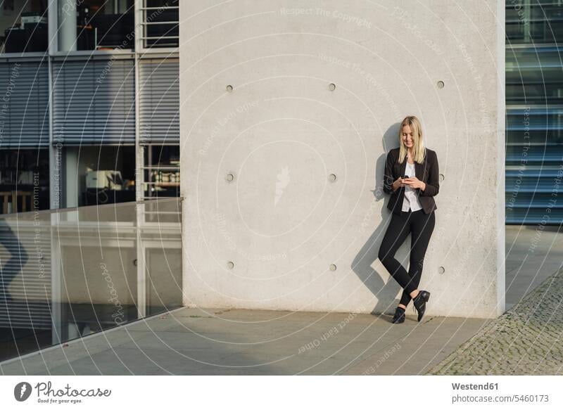 Blond businesswoman using smartphone leaning on wall human human being human beings humans person persons caucasian appearance caucasian ethnicity european 1