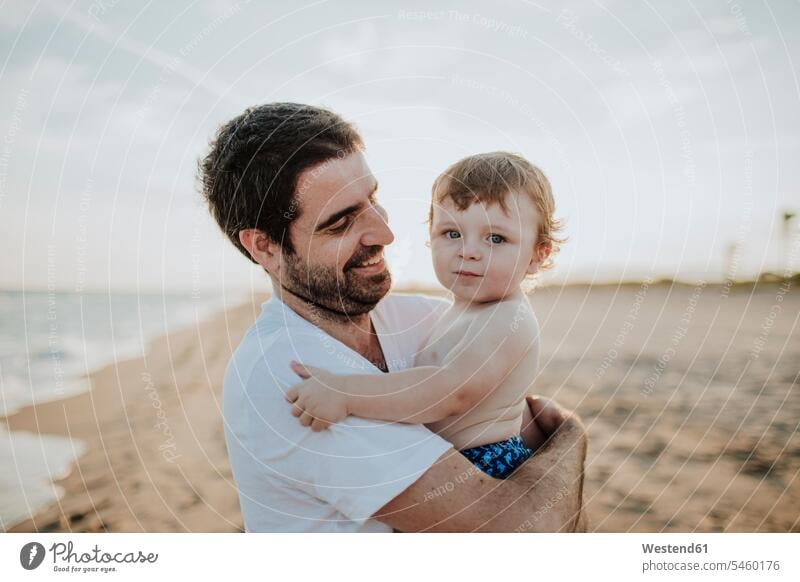 Smiling father carrying baby boy at beach during sunset color image colour image outdoors location shots outdoor shot outdoor shots sunsets sundown atmosphere