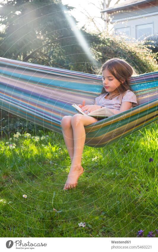 Little girl sitting on hammock in the garden reading a book females girls gardens Seated child children kid kids people persons human being humans human beings