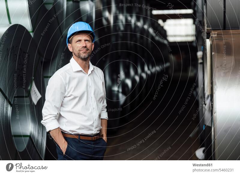 Businessman contemplating with hands in pockets standing by steel rolls in factory color image colour image indoors indoor shot indoor shots interior