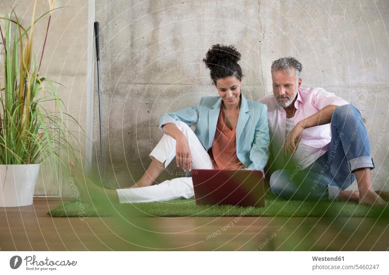 Casual businessman and businesswoman sitting on artificial turf in a loft sharing laptop Businessman Business man Businessmen Business men Laptop Computers