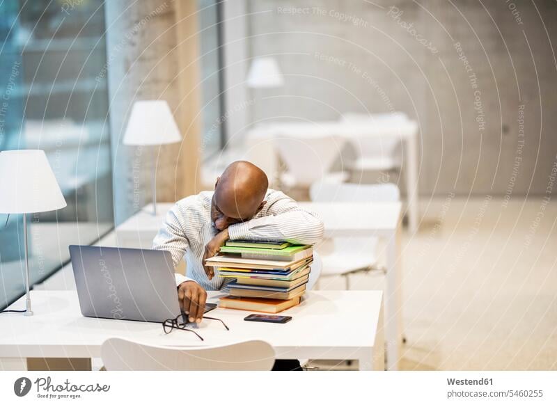 Exhausted mature man sitting at desk leaning on stack of books Exhaustion Weary exhausted Seated rested on stacks of books desks men males Table Tables Adults