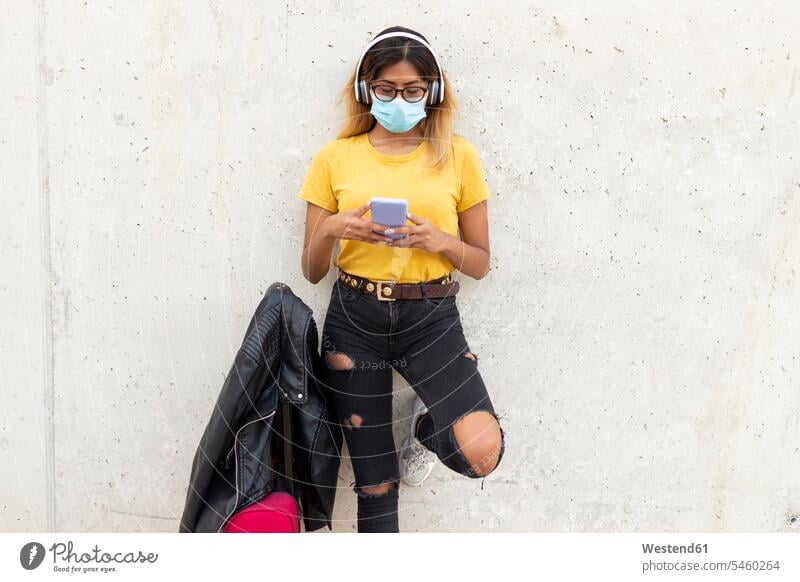 Young woman wearing protective mask while using smart phone by luggage against white wall at bus stop color image colour image casual clothing casual wear
