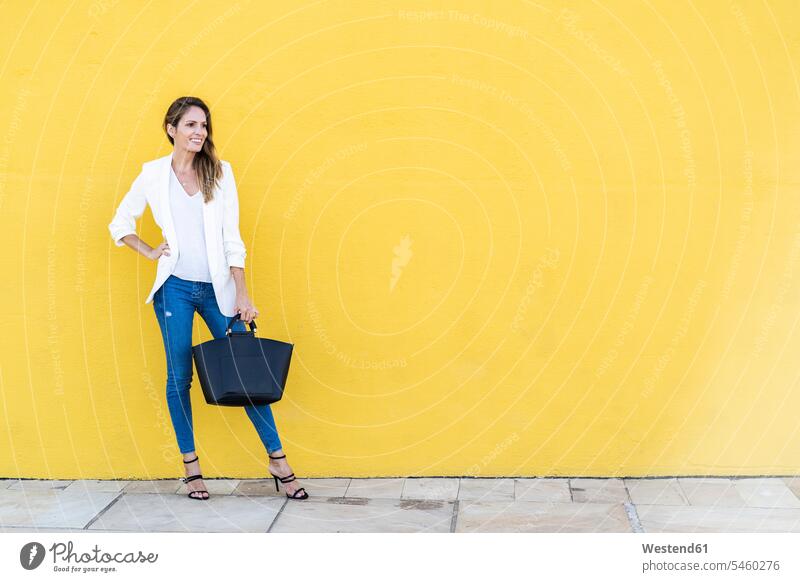 Smiling woman standing at a yellow wall holding a handbag human human being human beings humans person persons caucasian appearance caucasian ethnicity european