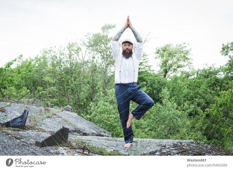 Bearded man wearing cap practicing tree pose on rock against trees color image colour image Austria outdoors location shots outdoor shot outdoor shots day