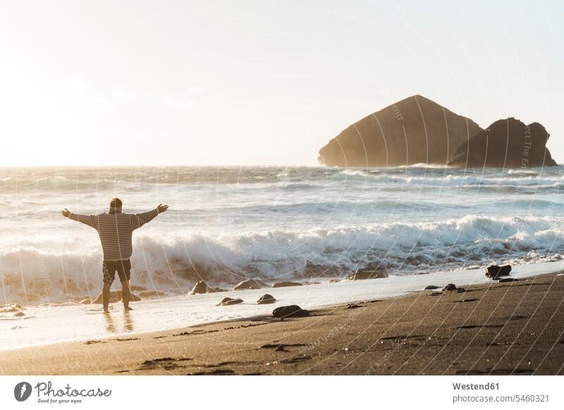 Man standing on the beach at sunset with outstretched arms, Sao Miguel Island, Azores, Portugal touristic tourists jumper sweater Sweaters relax relaxing