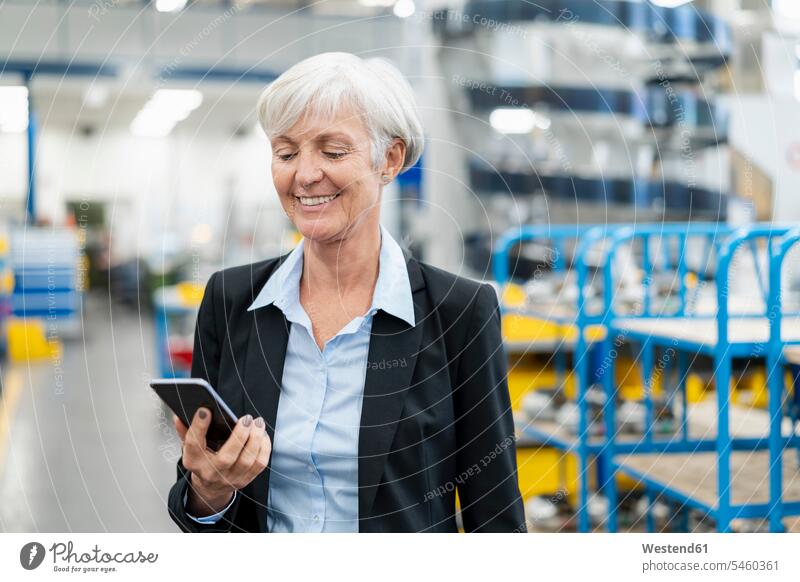 Smiling senior businesswoman looking at cell phone in a factory mobile phone mobiles mobile phones Cellphone cell phones eyeing factories businesswomen