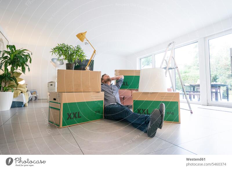 Man sitting on the floor in living room surrounded by cardboard boxes floors Seated man men males living rooms livingroom packing case packing cases Adults