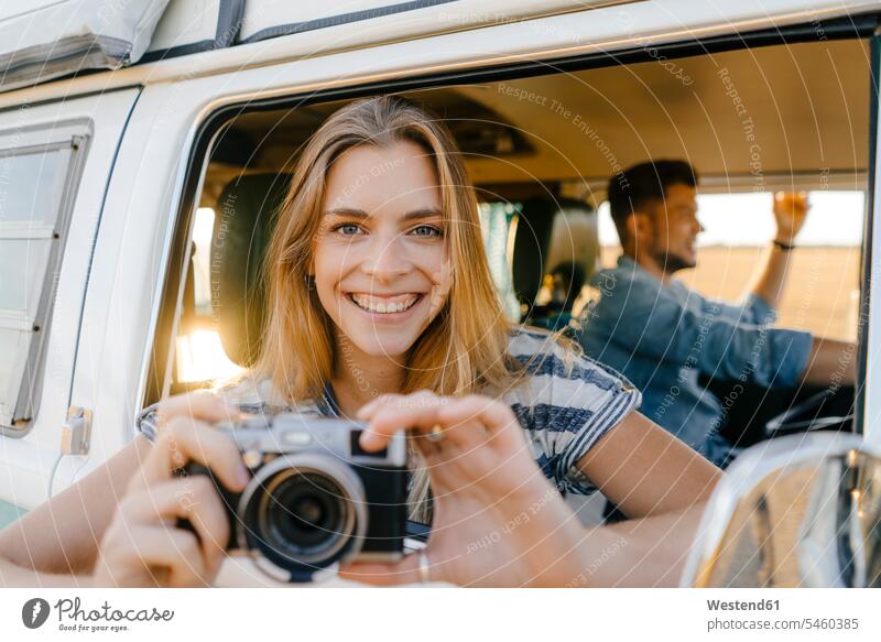 Portrait of happy woman with camera leaning out of window of a camper van with man driving portrait portraits couple twosomes partnership couples drive