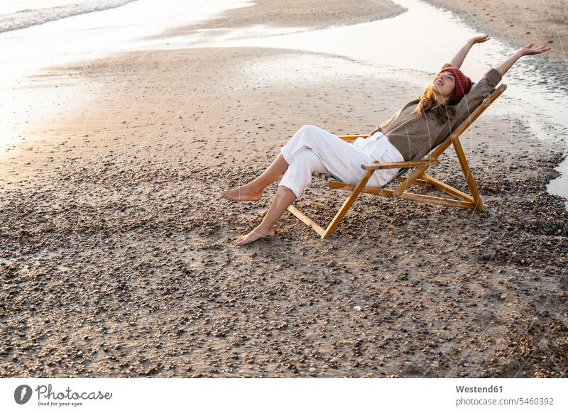Smiling young woman sitting with legs cross and arms raised on folding chair at beach during sunset color image colour image Netherlands Holland The Netherlands
