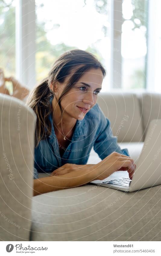 Smiling mature woman lying on couch at home using laptop couches settee settees sofa sofas computers Laptop Computer Laptop Computers laptops notebook smile