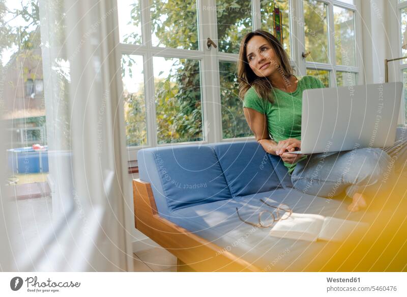 Smiling mature woman sitting on couch at home with laptop couches settee settees sofa sofas smile Seated computers Laptop Computer Laptop Computers laptops