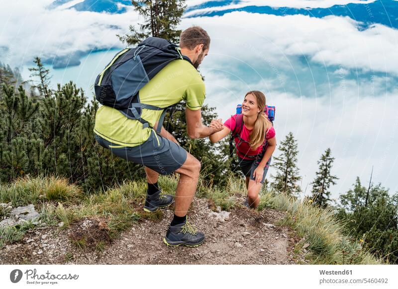 Young man helping girlfriend on a hiking trip in the mountains, Herzogstand, Bavaria, Germany touristic tourists back-pack back-packs backpacks rucksack