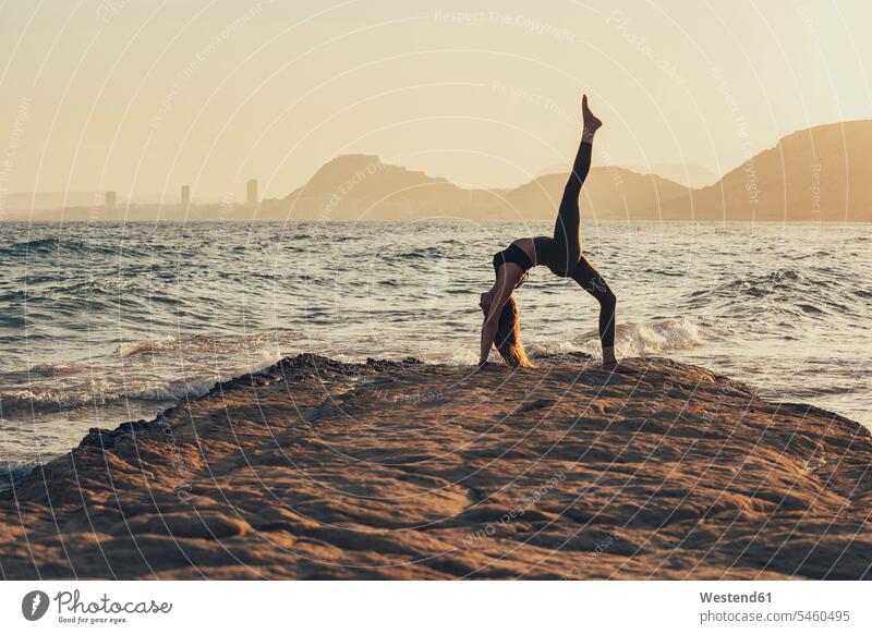 Mature woman practicing yoga at rocky beach in the evening, Urdhva Dhanurasana human human being human beings humans person persons caucasian appearance