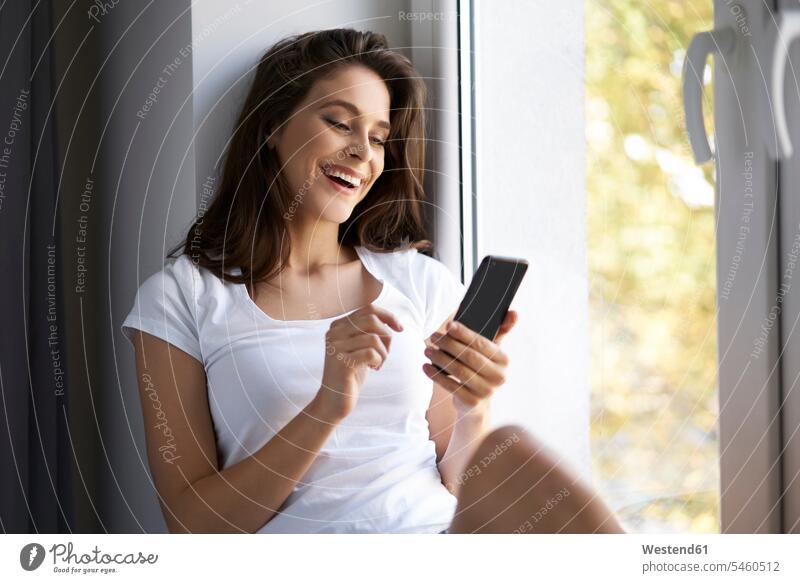 Young woman sitting on window sill, using smartphone Seated young women young woman home at home windowsill sills window sills Window Cill windowsills use