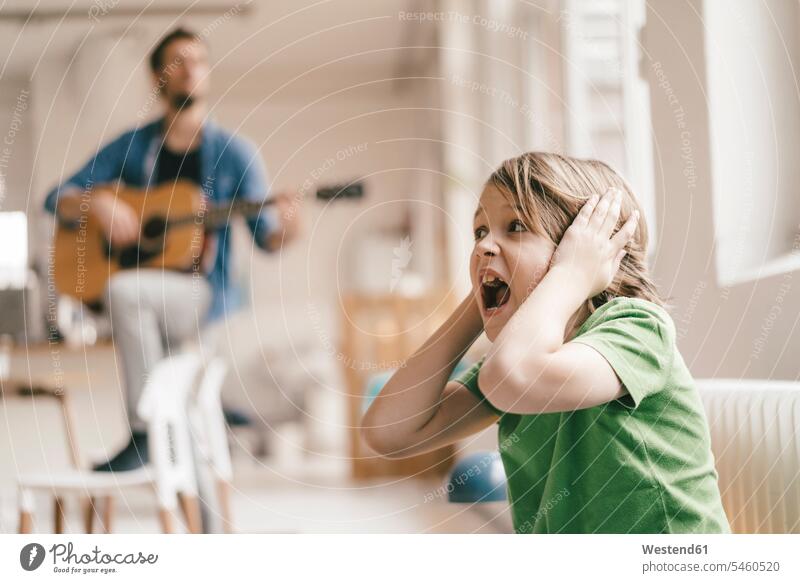 Horrified son covering his ears with father playing guitar at home making music playing music make music play music guitars sons manchild manchildren pa fathers