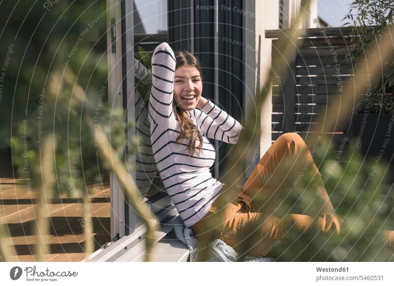 Portrait of laughing young woman sitting on terrace at home Germany comfortable hands behind head hands behind neck Hand Behind Head flat flats apartment