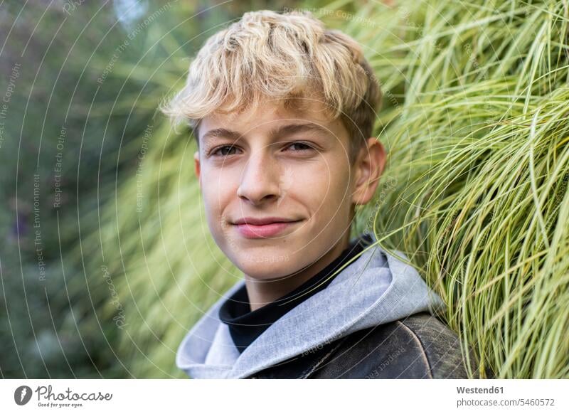 Confident blond teenage boy standing against plants color image colour image Germany outdoors location shots outdoor shot outdoor shots day daylight shot