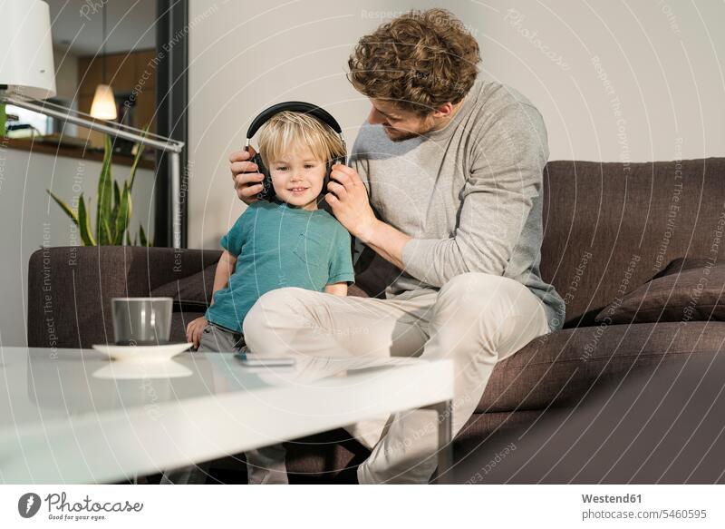 Father putting on headphones on son on couch at home sons manchild manchildren father pa fathers daddy dads papa headset settee sofa sofas couches settees