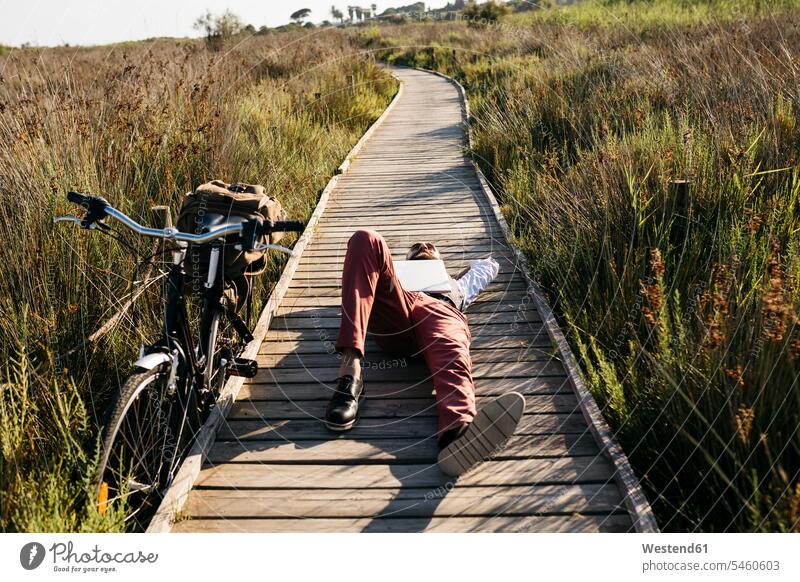 Well dressed man with laptop lying on a wooden walkway in the countryside next to a bike human human being human beings humans person persons