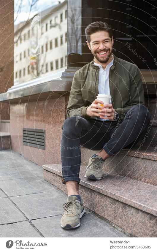 Portrait of laughing young man sitting on steps with coffee to go Seated delight enjoyment Pleasant pleasure free time leisure time funny having fun