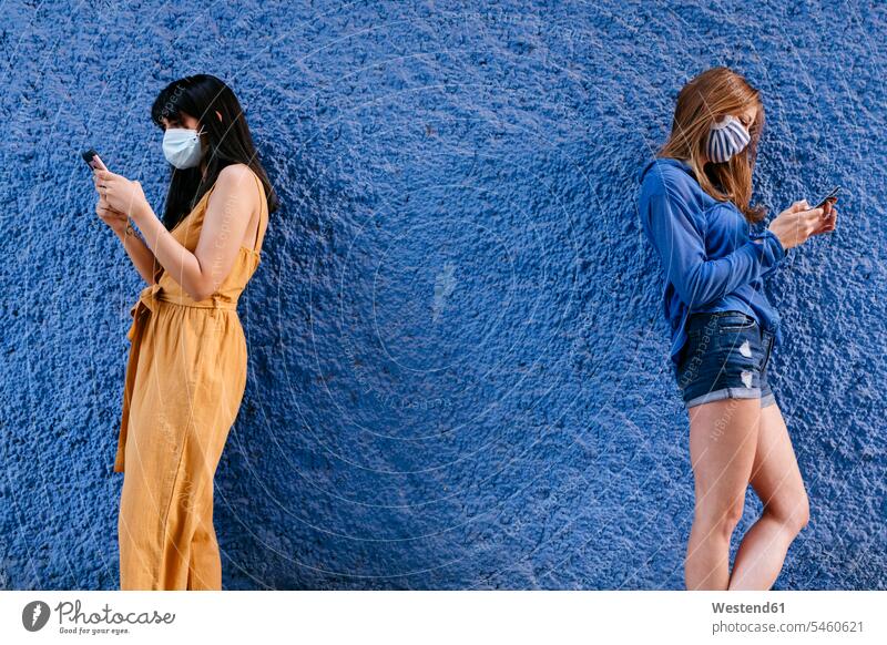 Female friends wearing masks using smart phones while standing by blue wall in city color image colour image Spain leisure activity leisure activities free time