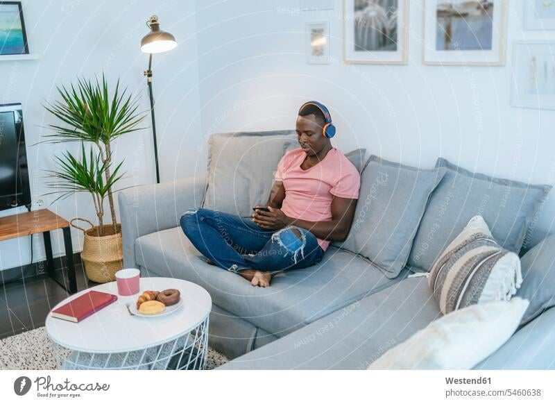 Young man with headphones and smartphone on couch at home human human being human beings humans person persons African black black ethnicity coloured 1
