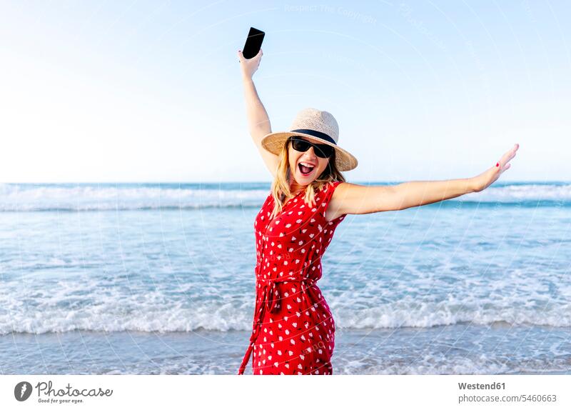 Smiling blond woman wearing red dress and hat and holding smartphone at the beach telecommunication phones telephone telephones cell phone cell phones Cellphone
