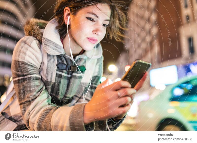 Spain, Madrid, young woman in the city at night using her smartphone and wearing earphones by night nite night photography ear phone ear phones Smartphone