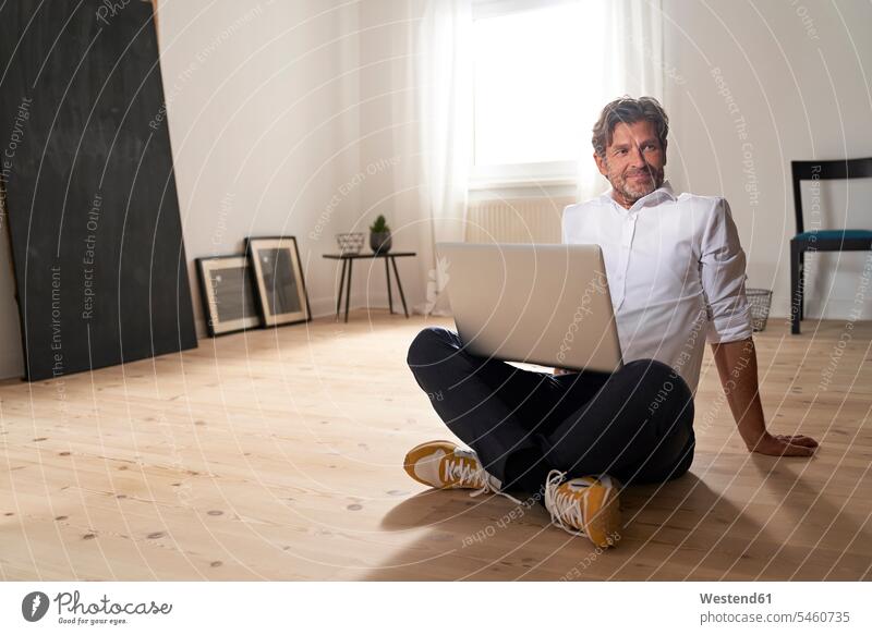Portrait of smiling mature man with laptop sitting on the floor at home frame frames picture frames Technological technologies The Internet e-mailing e-mails