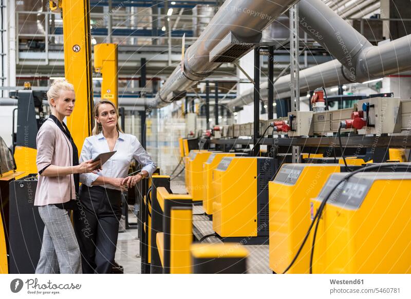 Two women with tablet looking at machine in factory shop floor factories woman females eyeing industrial hall factory hall industrial buildings digitizer