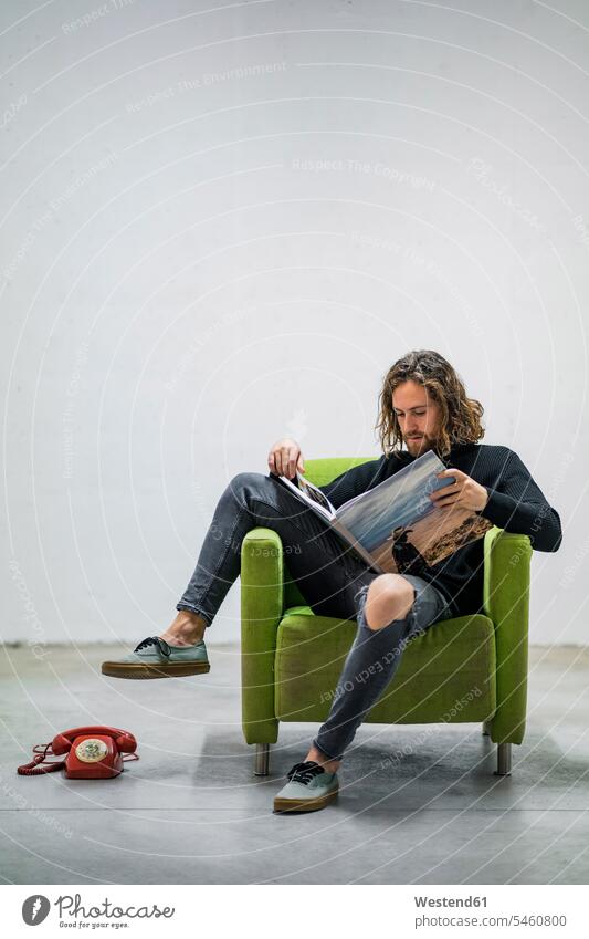 Young man reading book while sitting on armchair against wall at home color image colour image Spain leisure activity leisure activities free time leisure time
