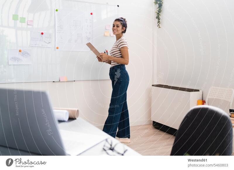 Smiling beautiful young female architect holding clipboard while standing by whiteboard in creative workplace color image colour image indoors indoor shot