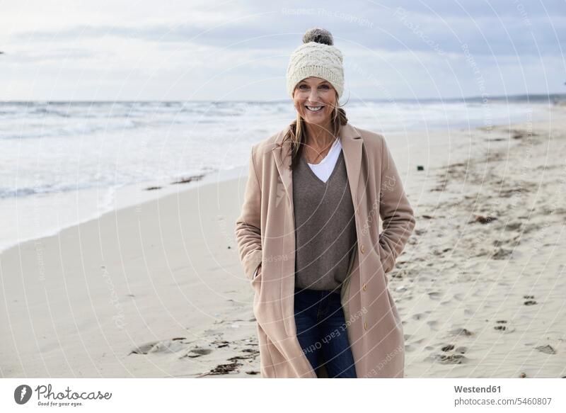 Spain, Menorca, portrait of smiling senior woman wearing bobble hat and coat on the beach in winter hibernal smile portraits bobble cap Coat Coats senior women