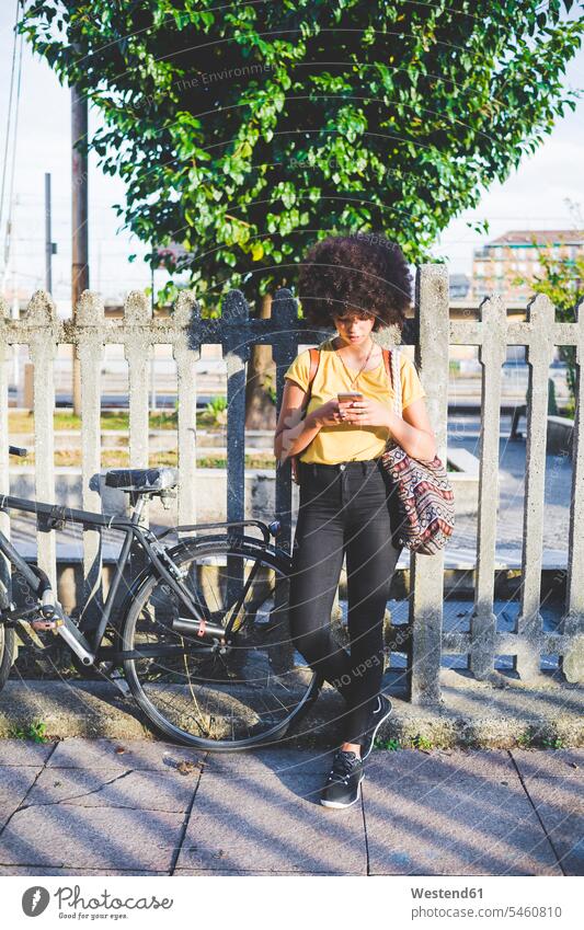 Young woman with afro hairdo using smartphone in the city human human being human beings humans person persons curl curled curls curly hair fences back-pack