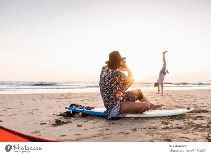 Young woman sitting on surfboard, taking pictures of young man, practicing handstands on the beach using use practice practise exercise exercising practising