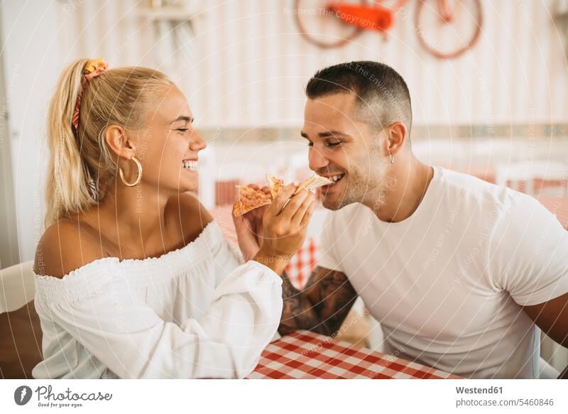 Cheerful young woman feeding pizza to boyfriend while sitting in restaurant color image colour image Spain leisure activity leisure activities free time