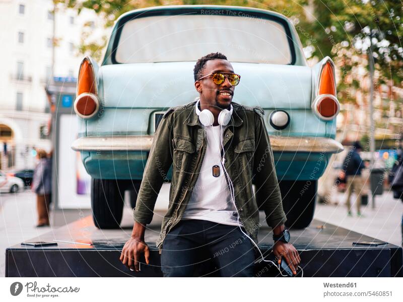 Young man wearing sunglasses with smart phone and headphones sitting against vintage car color image colour image Spain leisure activity leisure activities