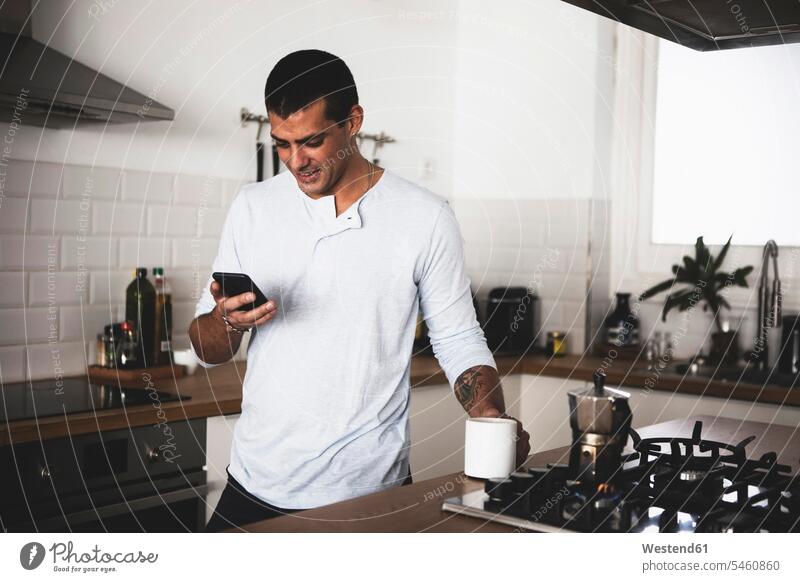 Smiling young man with cup of coffee using cell phone in kitchen at home Coffee smiling smile Coffee Cup Coffee Cups men males mobile phone mobiles