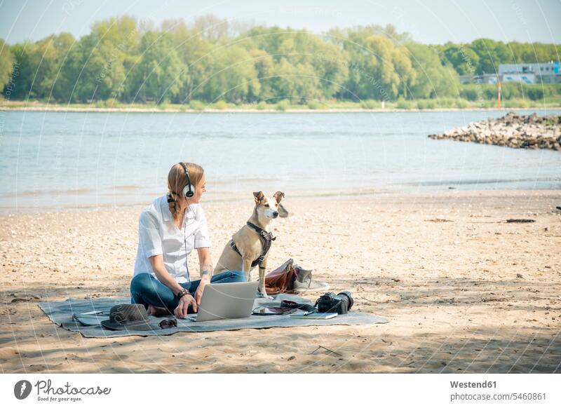 Woman sitting on blanket at a river with dog wearing headphones and using laptop Laptop Computers laptops notebook Blanket Blankets dogs Canine woman females