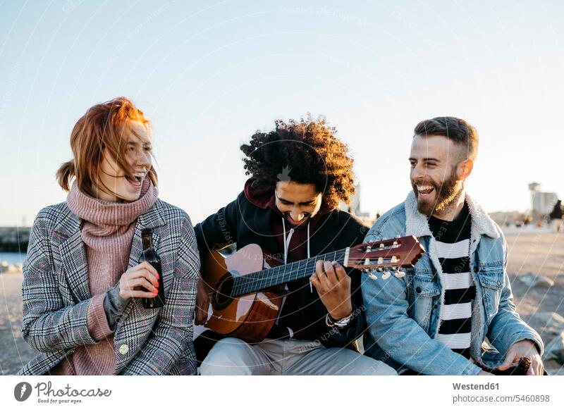 Three happy friends with guitar sitting outdoors at sunset happiness guitars Seated sunsets sundown friendship stringed instrument stringed instruments