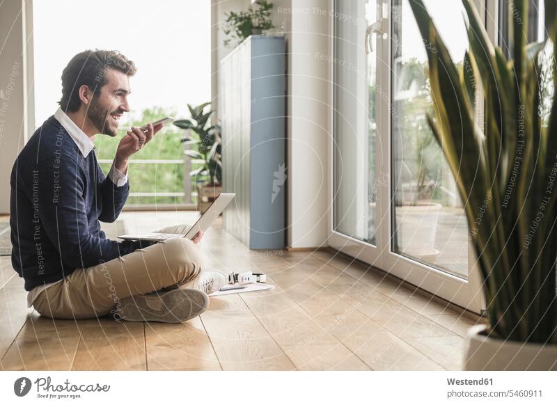 Young man sitting cross-legged in front of window, using laptop, talking on the phone speaking Foliage Plant Foliage Plants accessibility accessible