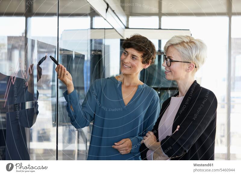 Two smiling businesswomen looking at chart on glass pane in office smile window windows graphic charts graphics eyeing offices office room office rooms